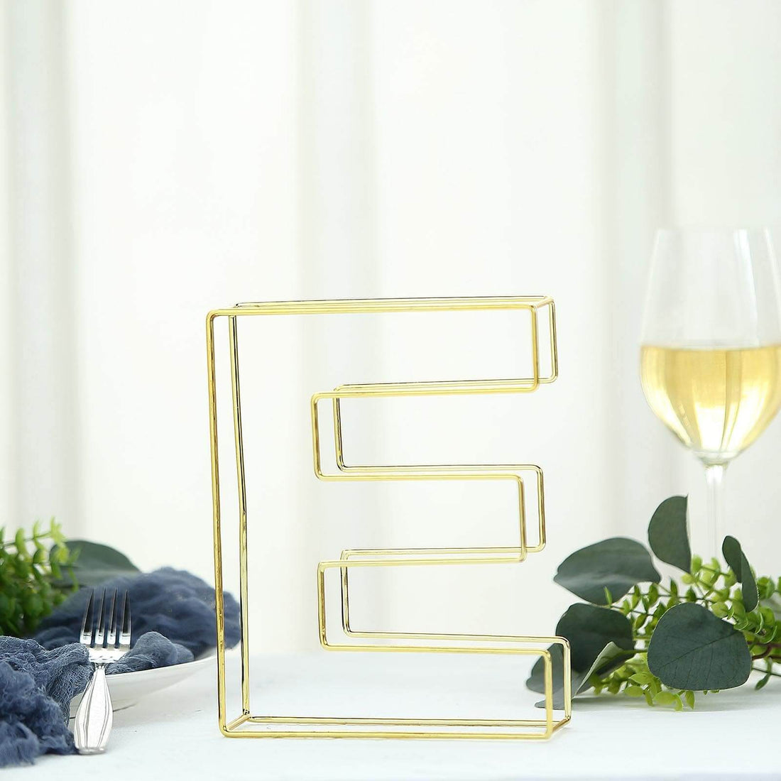 Gold wire letter "E" decorative piece on a table with greenery and a glass of wine.