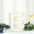 Gold wire letter "E" decorative piece on a table with greenery and a glass of wine.