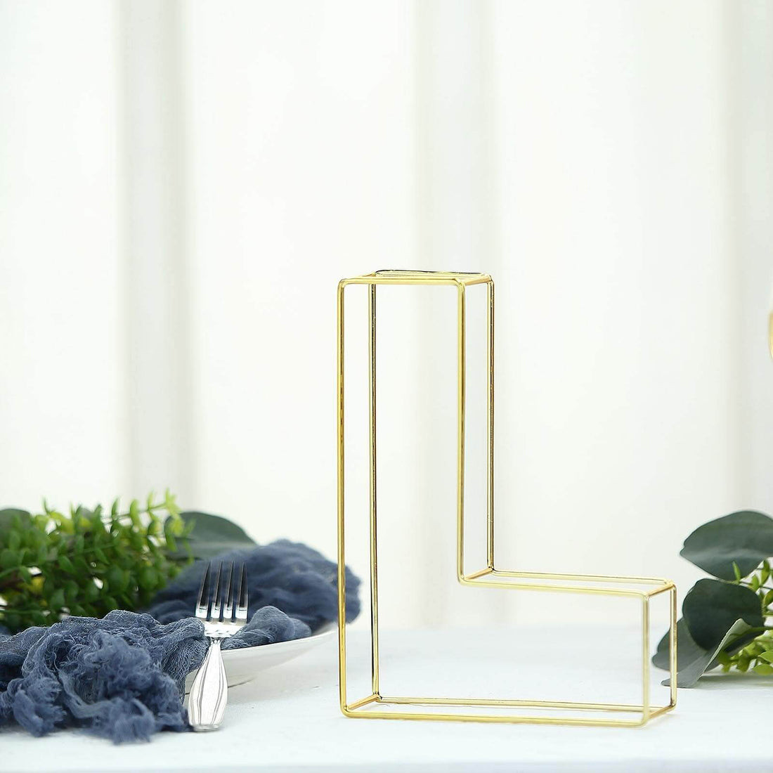 Gold geometric letter "L" decor on a table with greenery and fabric accents.