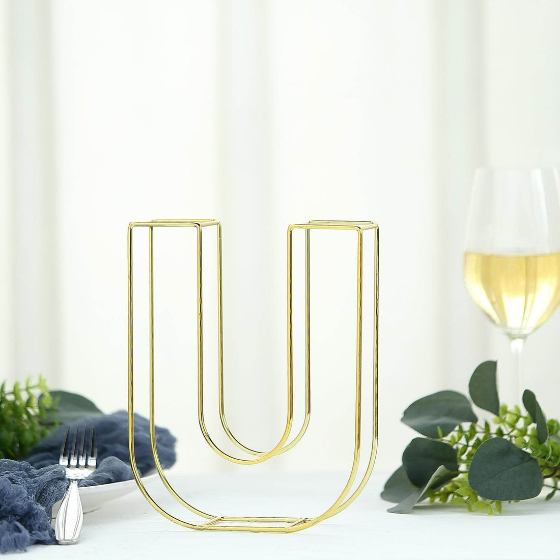 Gold U-shaped vase on a white table with greenery and wine glass.
