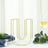 Gold U-shaped vase on a white table with greenery and wine glass.