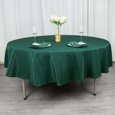Green round tablecloth draped over a table set for two with glasses and flowers.