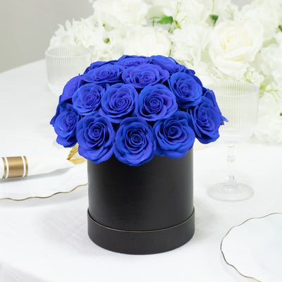 Blue roses in a black round box on a table with white flowers in the background.