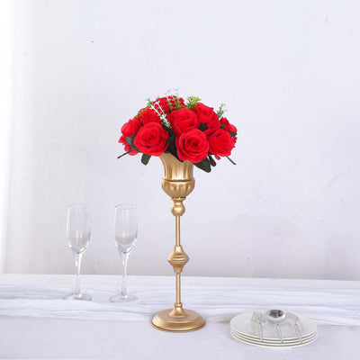 Gold vase with red roses and greenery on a table with glassware.