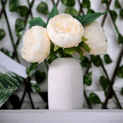 White vase with artificial peonies against a green backdrop.