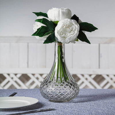 Clear glass vase with white peonies and green leaves on a table setting.