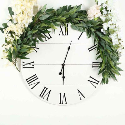 White round wall clock with Roman numerals surrounded by greenery and flowers.