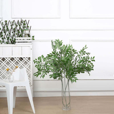 Artificial green leaf arrangement in a clear vase on a light wooden floor.