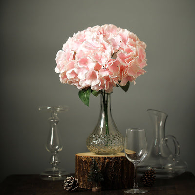 Pink hydrangea bouquet in glass vase on wooden stand with decorative elements.