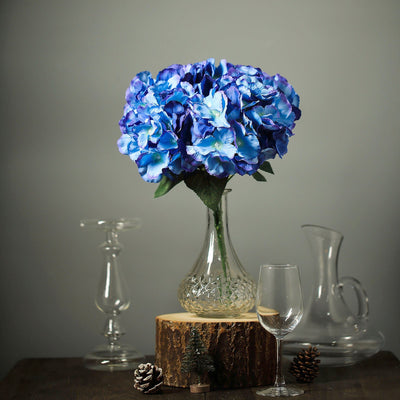 Blue hydrangea bouquet in a glass vase on a wooden stump with decorative glassware.