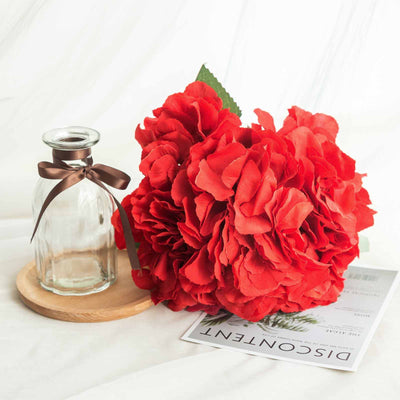 Red artificial flowers with a glass vase and wooden coaster.