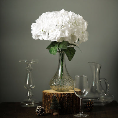 White hydrangea arrangement in a glass vase on a wooden slice with decorative elements.