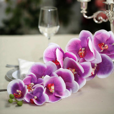 Purple orchid arrangement on a dining table with glassware and elegant decor.