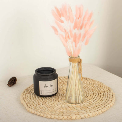 Pink dried flower arrangement in a glass vase with a black candle on a woven placemat.
