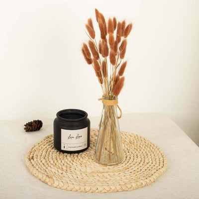 Decorative vase with dried flowers and candle on a woven mat.