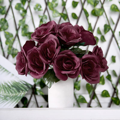 Burgundy artificial roses in a white vase against a greenery backdrop.