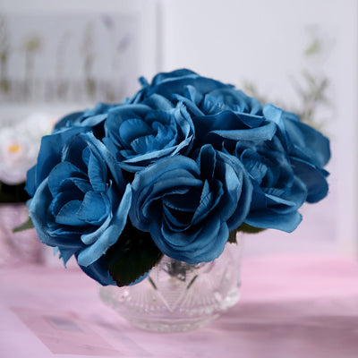 Blue silk rose bouquet in a glass vase on a pink surface.