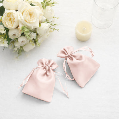 Two pink satin gift bags with drawstring, placed beside white roses and a candle.