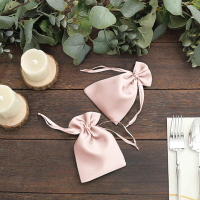 Two pink satin drawstring bags on a wooden table with greenery and candles.