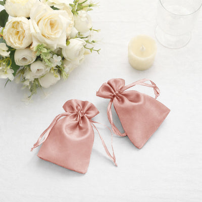 Two pink satin drawstring bags on a white table, surrounded by flowers and a candle.