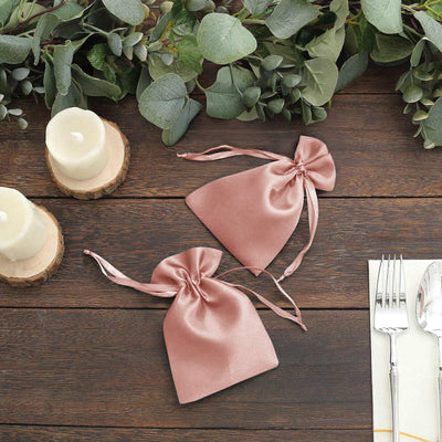 Two pink satin drawstring bags on a wooden table with greenery and candles.