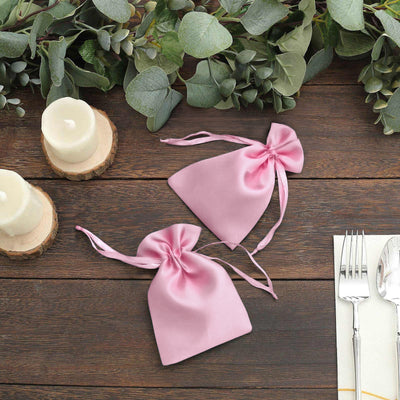 Pink satin favor bags on a rustic wooden table with greenery and candles.