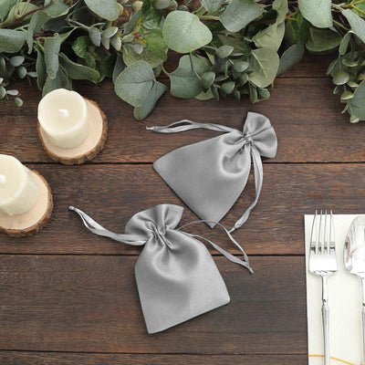 Two elegant gray satin drawstring bags on a wooden table with greenery and candles.