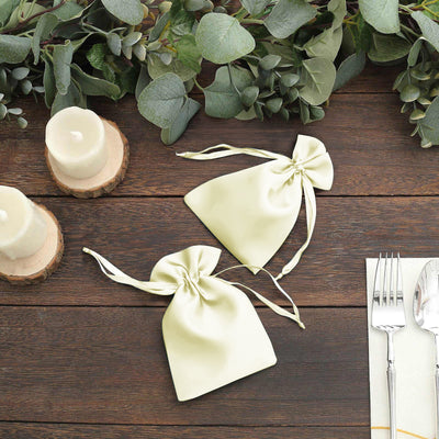 Two light green drawstring gift bags on a wooden table with candles and greenery.