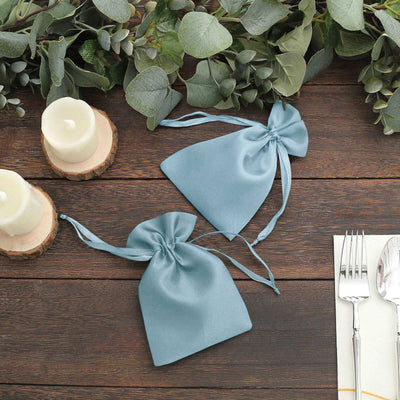Two blue satin gift bags on a wooden table with greenery and candles.