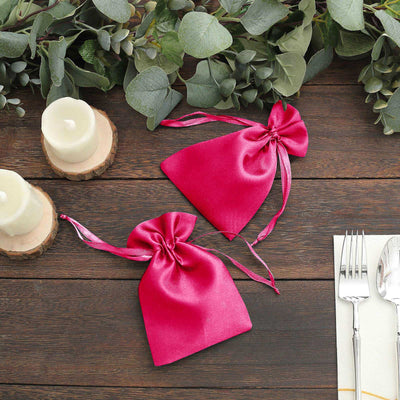 Two pink satin drawstring bags on a rustic wooden table with candles and greenery.