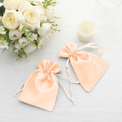 Two peach satin drawstring bags on a table with flowers and a candle.