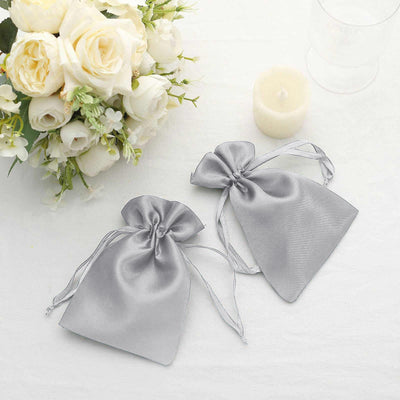 Silver satin drawstring bags on a table with flowers and a candle.