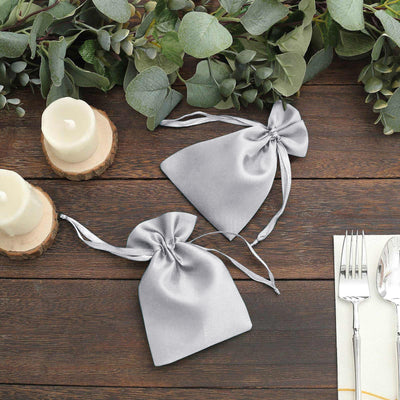 Silver satin drawstring bags on a wooden table with candles and greenery.