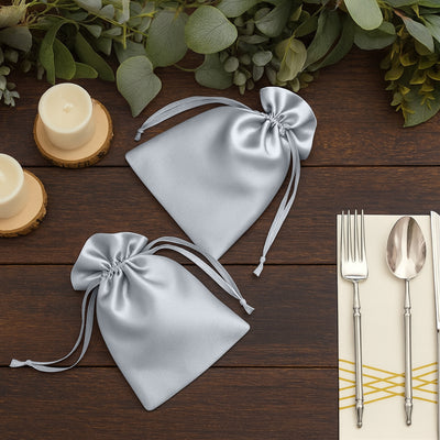 Silver satin drawstring bags on a wooden table with candles and cutlery.