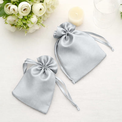 Silver satin drawstring bags on a soft surface with flowers and a candle.