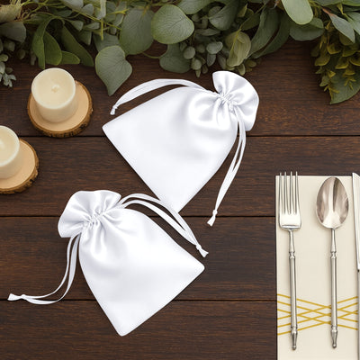 White satin favor bags on a wooden table with candles and silverware.