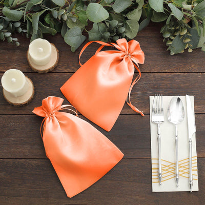 Orange satin gift bags on a wooden table with utensils and candles.