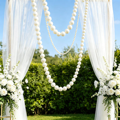 Elegant white wedding arch with drapes, pearl garlands, and floral arrangements outdoors
