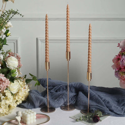 Elegant twisted candles in gold holders with floral arrangement backdrop.
