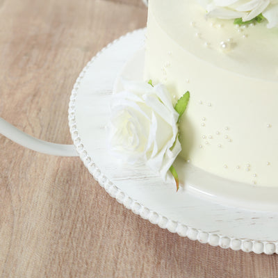 White frosted cake adorned with white roses and pearls on a decorative platter.