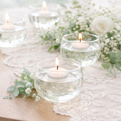 Floating candles in glass bowls with greenery on a lace table setting.