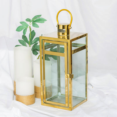 Gold metal and glass lantern with handle, surrounded by candles and greenery.