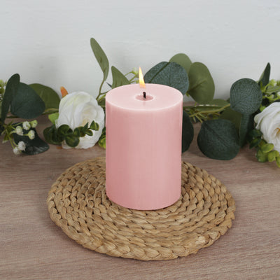 Pink pillar candle on a woven placemat with greenery and flowers around it.