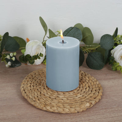Blue pillar candle on a woven mat with greenery and flowers in the background.