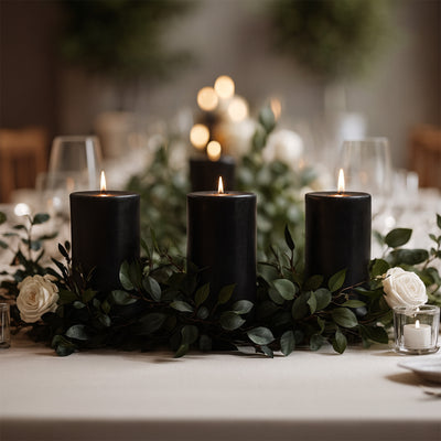 Black pillar candles with greenery and white roses on a dining table setting.