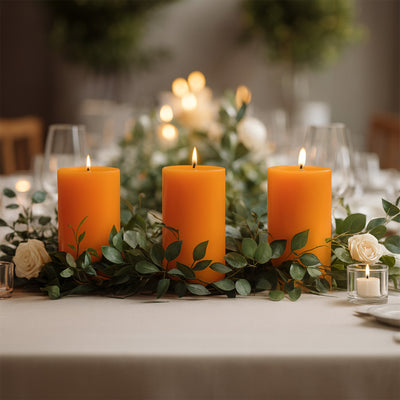Three orange candles surrounded by greenery and white roses on a dining table.
