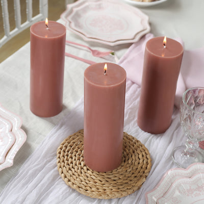 Three pink pillar candles on a woven placemat with elegant dinnerware in the background.