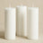 Three white pillar candles in varying heights on a soft beige background.