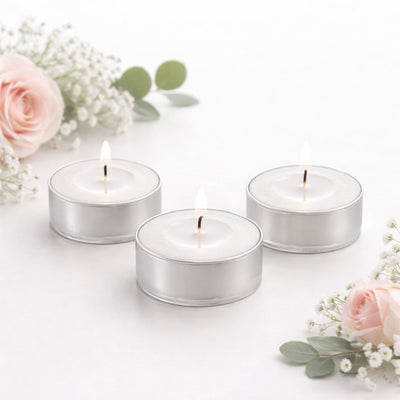 Three lit silver tealight candles surrounded by pink roses and greenery.