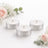 Three lit silver tealight candles surrounded by pink roses and greenery.